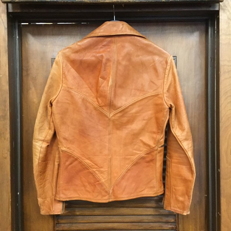 May include: A brown leather jacket with a V-shaped back panel and a collar. The jacket has long sleeves and a zipper closure.