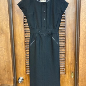 Puede incluir: Un vestido midi negro con cuello redondo, mangas cortas y cintur&oacute;n. El vestido tiene cierre de botones en la parte delantera y un ribete de cuero.