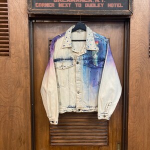 May include: A light blue denim jacket with a tie-dye effect in shades of purple and blue. The jacket is decorated with rhinestones and has a faded look.