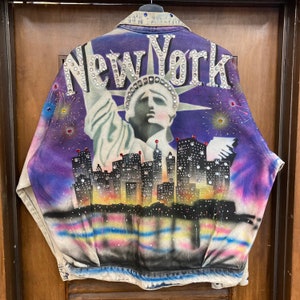 May include: A denim jacket with a painted design of the Statue of Liberty and the New York City skyline. The jacket is decorated with rhinestones and has a purple, blue, and pink color scheme.