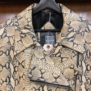 Puede incluir: Una chaqueta de cuero beige con un estampado de piel de serpiente negra. La chaqueta tiene una cremallera y un cuello. Una etiqueta con el texto "Michael  H. Hart North Beach" est&aacute; unida a la chaqueta.