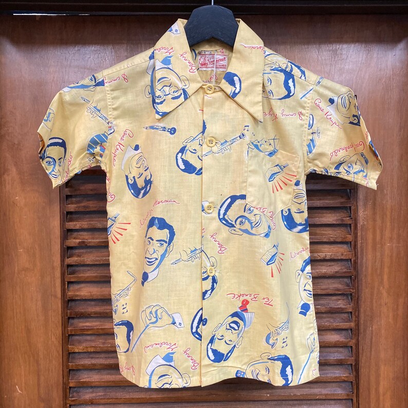 May include: A yellow short-sleeved button-down shirt with a pattern of cartoon faces and musical instruments. The shirt has a collar and a pocket on the left side. The text on the shirt includes "Tex Beneke", "Benny Goodman", "Gene Krupa", and "Harry James".