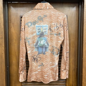 May include: A brown long-sleeve shirt with a brick pattern and a graphic print of a person standing in front of a slot machine. The graphic has the text "BIG 817" and "1234567890".