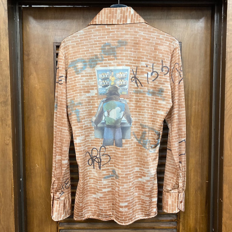 May include: A brown long-sleeve shirt with a brick pattern and a graphic print of a person standing in front of a slot machine. The graphic has the text "BIG 817" and "1234567890".