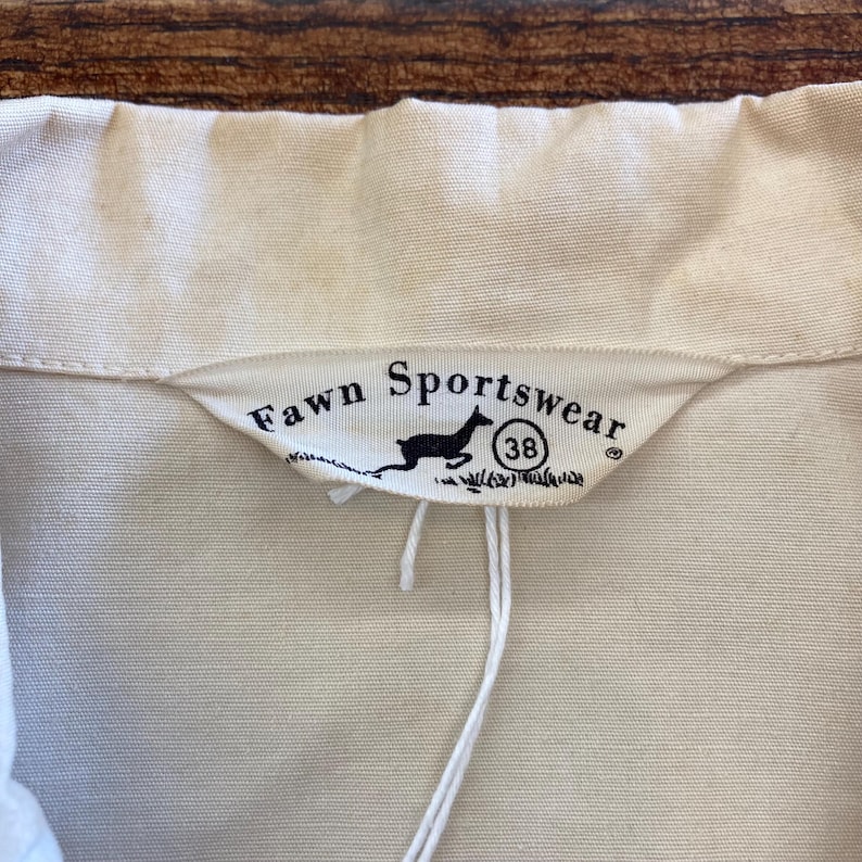 Puede incluir: Una etiqueta blanca con texto negro que dice "Fawn Sportswear" y una peque&ntilde;a silueta negra de un ciervo corriendo. La etiqueta tambi&eacute;n tiene un c&iacute;rculo blanco con el n&uacute;mero "38" dentro.