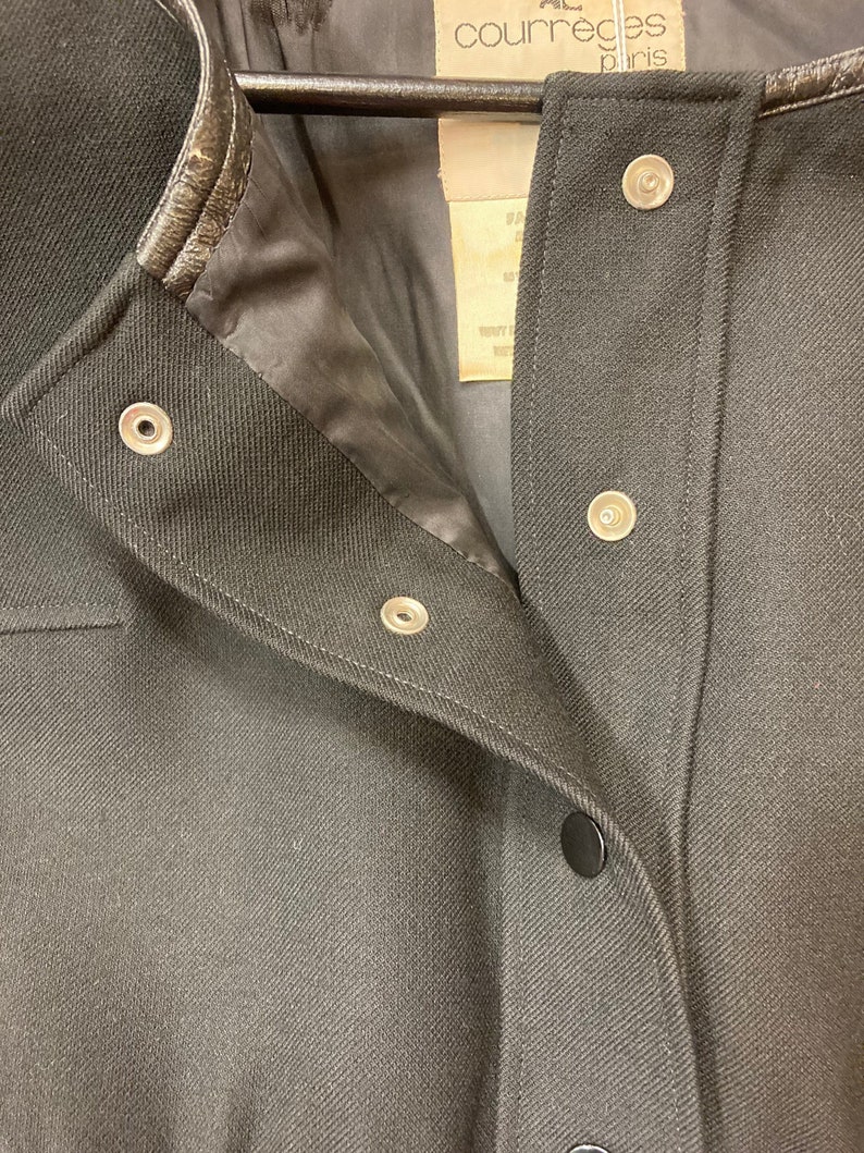 Puede incluir: Chaqueta negra con cuello de cuero y cierres a presi&oacute;n. La chaqueta tiene la etiqueta Courr&egrave;ges Paris en la parte delantera.