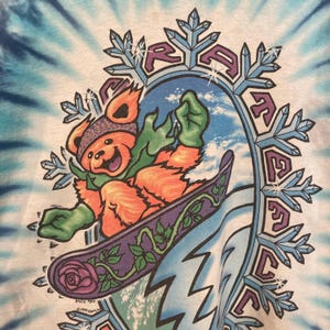 Puede incluir: Camiseta te&ntilde;ida con un oso de dibujos animados practicando snowboard. El oso lleva gorro y guantes, con una tabla de snowboard morada decorada con rosas y vides. El dise&ntilde;o incluye la palabra "Grateful" y detalles de copos de nieve.