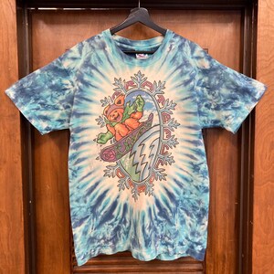 Puede incluir: Camiseta azul te&ntilde;ida con un dise&ntilde;o de un oso esquiando dentro de un marco con forma de copo de nieve. El oso lleva ropa naranja y un gorro. La camiseta tiene cuello redondo y mangas cortas.