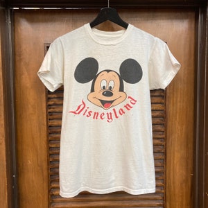 Peut inclure: T-shirt blanc avec un motif vintage de Mickey Mouse et le texte "Disneyland" en rouge.