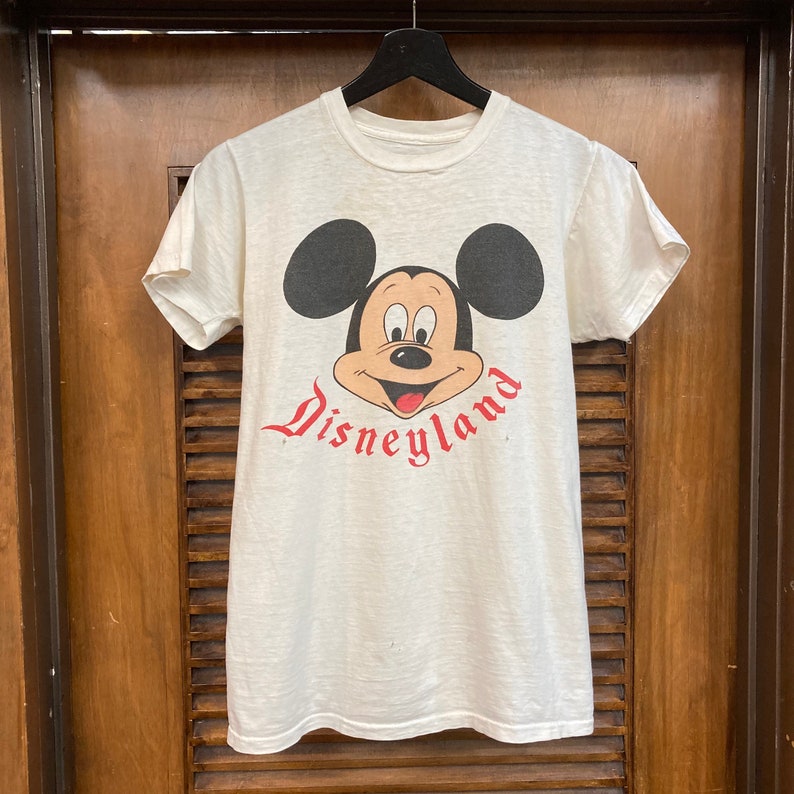 Peut inclure: T-shirt blanc avec un motif vintage de Mickey Mouse et le texte "Disneyland" en rouge.