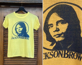 Puede incluir: Camiseta vintage amarilla con una imagen en negro y azul de Jackson Browne. La imagen está en un círculo con el texto "Jackson Browne" debajo.