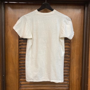 Peut inclure: T-shirt blanc &agrave; manches courtes suspendu &agrave; un cintre en bois sur un fond en bois.