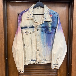 May include: A light blue denim jacket with a faded ombre effect, featuring a purple and blue design on the back. The jacket is decorated with rhinestones.