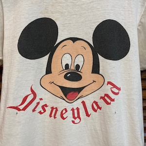 Peut inclure: T-shirt blanc avec un logo "Disneyland" rouge et une image de dessin anim&eacute; de Mickey Mouse avec des oreilles noires et un visage brun.
