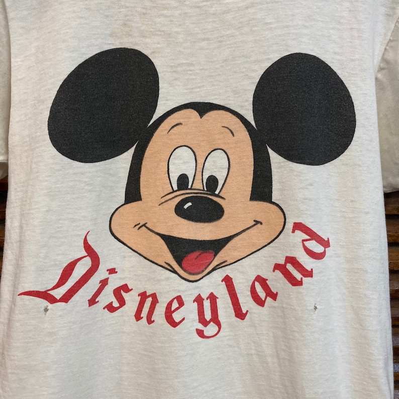 Peut inclure: T-shirt blanc avec un logo "Disneyland" rouge et une image de dessin anim&eacute; de Mickey Mouse avec des oreilles noires et un visage brun.