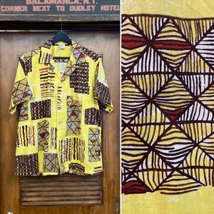 May include: Yellow short-sleeve button-down shirt with a geometric pattern in brown, white, and red. The shirt is hanging on a black hanger against a wooden door. The pattern is repeated on the right side of the image.