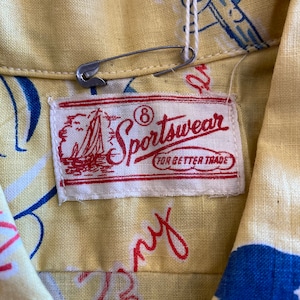 May include: A white label with red text that reads "Sportswear for Better Trade" with a small sailboat graphic and the number 8 circled. The label is attached to a yellow fabric shirt with a blue and white pattern.