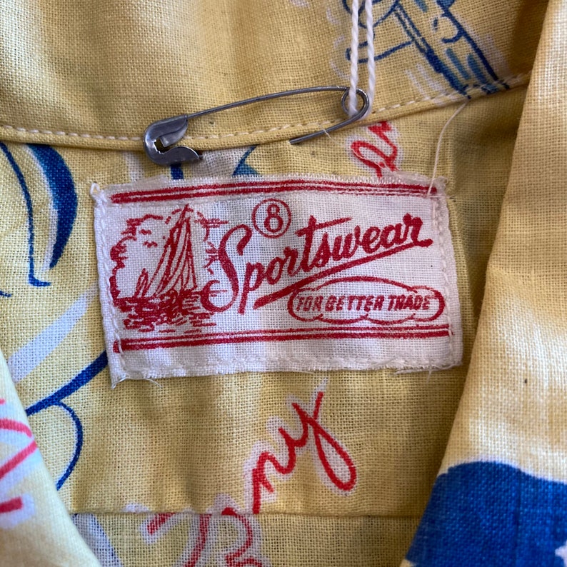 May include: A white label with red text that reads "Sportswear for Better Trade" with a small sailboat graphic and the number 8 circled. The label is attached to a yellow fabric shirt with a blue and white pattern.