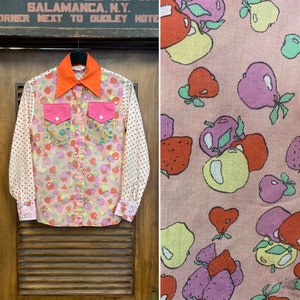 Könnte beinhalten: Ein rosa-weißes langärmliges Hemd mit Knöpfen und einem Fruchtmuster. Das Hemd hat einen roten Kragen und zwei rosa Taschen mit Strasssteinen. Die Ärmel haben weiße Punkte.