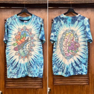 Puede incluir: Camiseta te&ntilde;ida con un dise&ntilde;o colorido. La parte delantera presenta un oso practicando snowboard a trav&eacute;s de un copo de nieve, y la parte trasera tiene un dise&ntilde;o similar con un personaje diferente. La camiseta es azul y blanca.