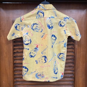 May include: A yellow short-sleeved button-down shirt with a repeating pattern of cartoon faces and musical instruments. The faces are of famous jazz musicians, including Benny Goodman, Count Basie, and Gene Krupa. The text on the shirt includes the names of the musicians and the names of their bands.