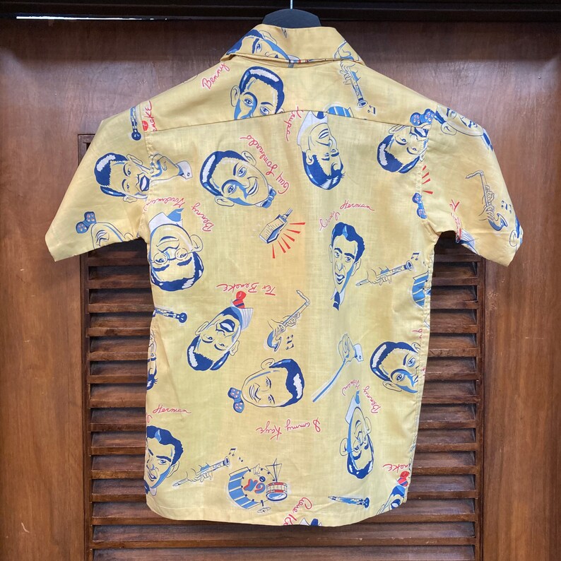 May include: A yellow short-sleeved button-down shirt with a repeating pattern of cartoon faces and musical instruments. The faces are of famous jazz musicians, including Benny Goodman, Count Basie, and Gene Krupa. The text on the shirt includes the names of the musicians and the names of their bands.
