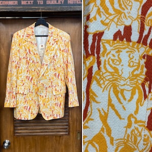 May include: A yellow, orange, and white patterned blazer with a two-button closure and two front pockets. The pattern features a tiger face and leaf design. The blazer is hanging on a black hanger against a wooden door.