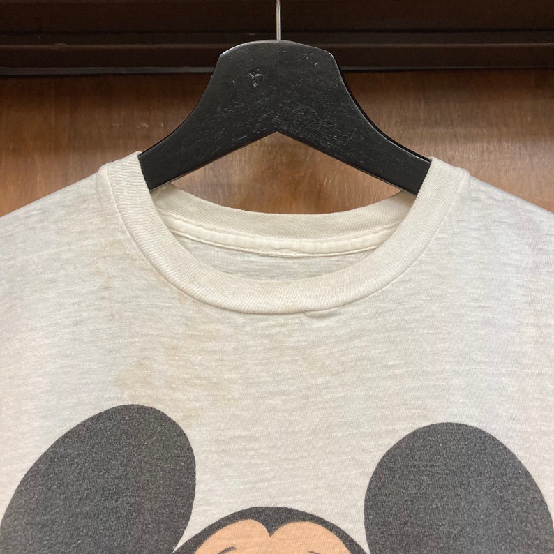 Peut inclure: Un t-shirt blanc avec une image en noir et blanc de Mickey Mouse. Le t-shirt est suspendu &agrave; un cintre noir.