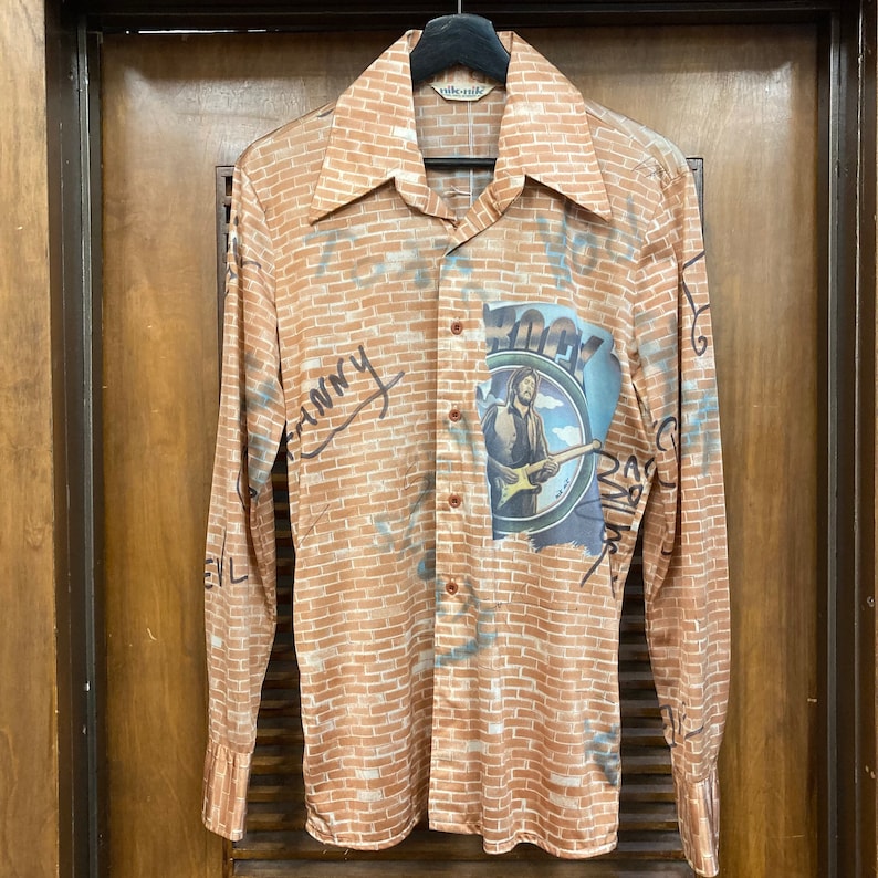 May include: A long-sleeved button-up shirt with a brick pattern and a graphic of a man playing a guitar. The shirt has the word "ROCKY" written on it.