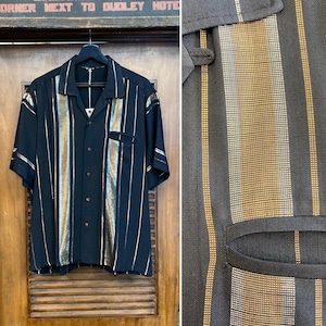 Puede incluir: Camisa negra de manga corta con rayas verticales doradas y plateadas. La camisa tiene un cuello clásico y un bolsillo en el pecho. Cuelga de una percha negra. Fabricada con tejido ligero.