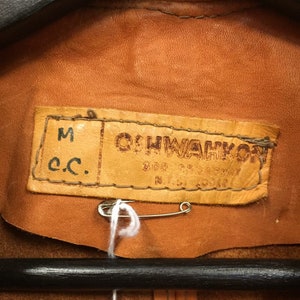 May include: Close-up of a brown leather label with the text "OHWAKON" and "M c.c." The label is attached to a brown leather jacket.