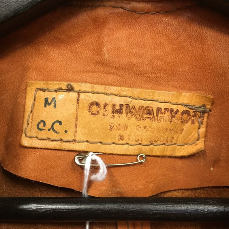 May include: Close-up of a brown leather label with the text "OHWAKON" and "M c.c." The label is attached to a brown leather jacket.