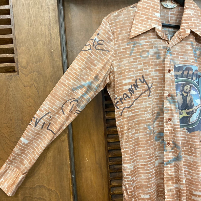May include: A long-sleeved shirt with a brick pattern and graffiti-style writing. The writing includes the words "Franny" and "Evil". The shirt also features a graphic of a person playing a guitar.