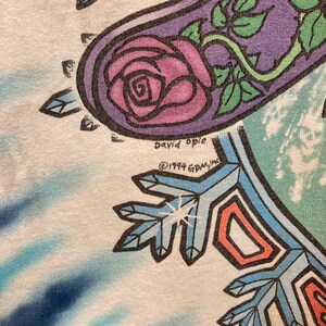 Puede incluir: Primer plano de una camiseta blanca con un dise&ntilde;o gr&aacute;fico colorido. El dise&ntilde;o incluye una rosa morada, vides verdes y formas geom&eacute;tricas en azul, naranja y blanco. Tambi&eacute;n se ve el texto "David Opie" y "&copy;1994 GDM, Inc".