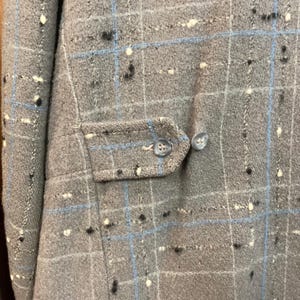 May include: A close-up of a grey plaid fabric with blue and white accents. The fabric has a textured surface and is likely a wool blend. The fabric is used for a jacket or coat.