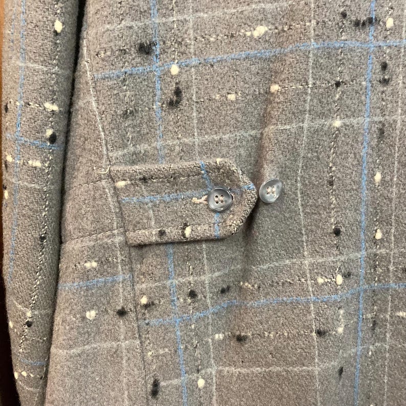 May include: A close-up of a grey plaid fabric with blue and white accents. The fabric has a textured surface and is likely a wool blend. The fabric is used for a jacket or coat.