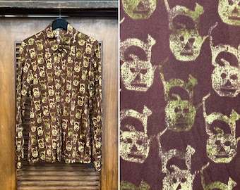 May include: A brown long-sleeved shirt with a repeating pattern of yellow stylized faces. The shirt has a collared neckline and button closure.
