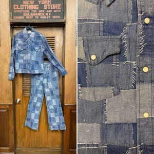 May include: A blue denim jacket and pants set made of patchwork fabric. The jacket has a button-up front and a pointed collar. The pants are flared and have a straight leg. The fabric is a mix of light and dark blue denim.