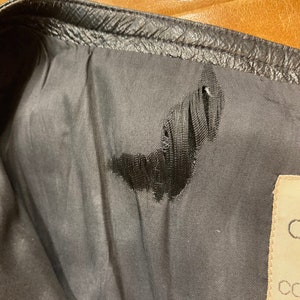 Puede incluir: Primer plano del forro de una chaqueta de cuero negra con un peque&ntilde;o desgarro en la tela. El forro es de color gris oscuro y el desgarro tiene la forma de una luna creciente. Una etiqueta blanca con texto negro es visible en la esquina inferior derecha de la imagen.