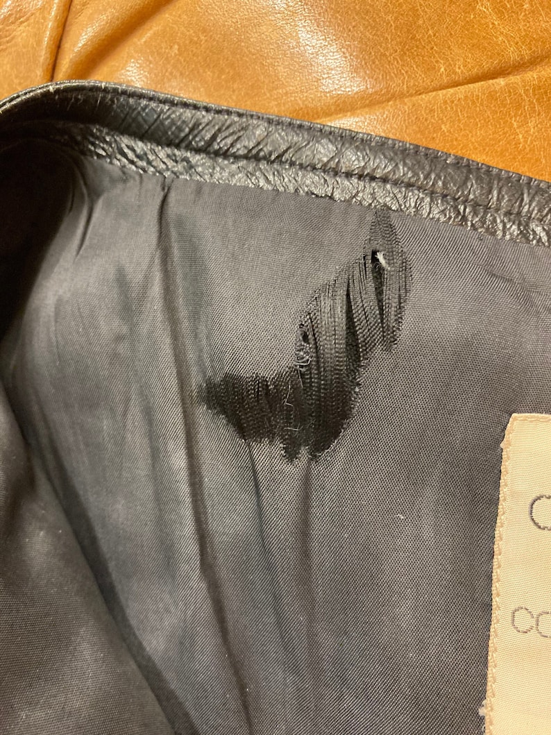 Puede incluir: Primer plano del forro de una chaqueta de cuero negra con un peque&ntilde;o desgarro en la tela. El forro es de color gris oscuro y el desgarro tiene la forma de una luna creciente. Una etiqueta blanca con texto negro es visible en la esquina inferior derecha de la imagen.