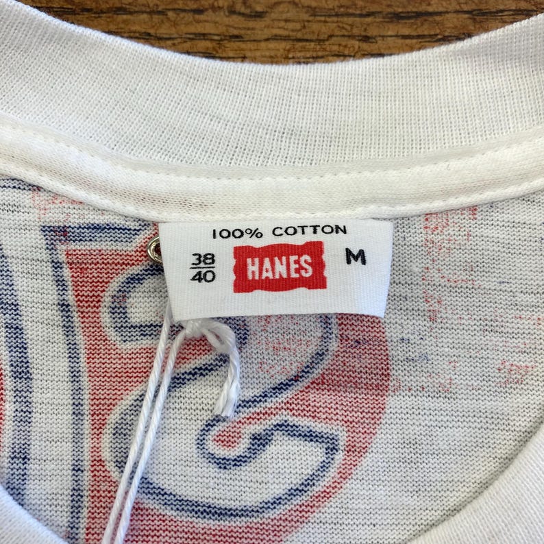 Peut inclure: Un t-shirt blanc avec un imprim&eacute; graphique rouge et bleu visible &agrave; travers le tissu. L'&eacute;tiquette int&eacute;rieure au col indique 100% COTTON, 38/40, HANES et M. Une &eacute;tiquette en ficelle blanche est attach&eacute;e. Le tissu pr&eacute;sente un col rond blanc c&ocirc;tel&eacute;.