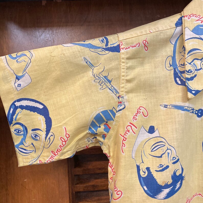 May include: A yellow short-sleeved shirt with a blue and white patterned design. The shirt features a repeating pattern of men's faces and musical instruments. The text "Gova Luhr" and "& Company" is printed on the shirt.