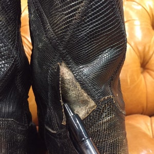 Pu&ograve; includere: Primo piano di un paio di stivali da cowboy in pelle nera con motivo a pelle di serpente. Gli stivali hanno un piccolo strappo sul lato di uno stivale.