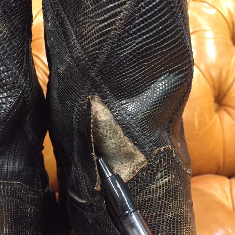 Pu&ograve; includere: Primo piano di un paio di stivali da cowboy in pelle nera con motivo a pelle di serpente. Gli stivali hanno un piccolo strappo sul lato di uno stivale.