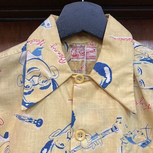 May include: A yellow button-down shirt with a cartoon print of a man with glasses and a musical note. The shirt has a label that reads "Sportwear" and the words "Downtown" and "Journey" are printed on the collar.
