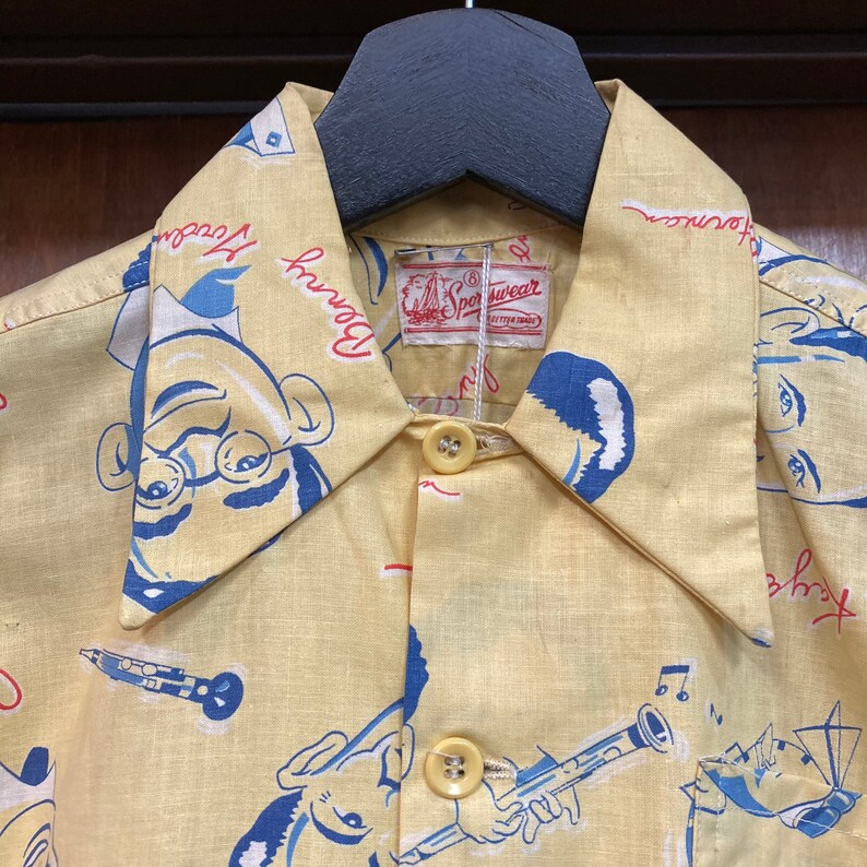 May include: A yellow button-down shirt with a cartoon print of a man with glasses and a musical note. The shirt has a label that reads "Sportwear" and the words "Downtown" and "Journey" are printed on the collar.