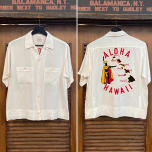 May include: White short-sleeve shirt with two chest pockets. The back of the shirt features a colorful "Aloha Hawaii" design with a figure and island map. The shirt is hanging on a wooden door.