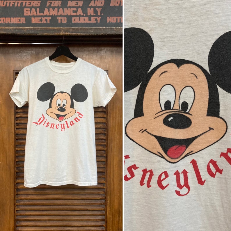 Peut inclure: T-shirt blanc avec un motif vintage de Mickey Mouse et le texte "Disneyland" en rouge.