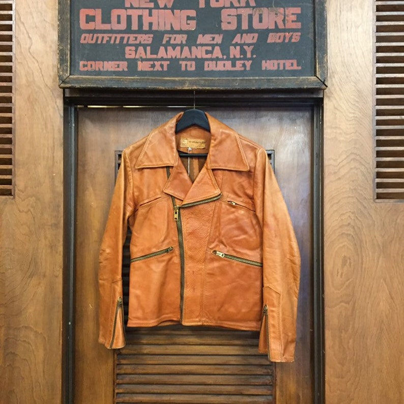 May include: A brown leather motorcycle jacket with a zipper closure and multiple pockets. The jacket is hanging on a hanger against a wooden background.