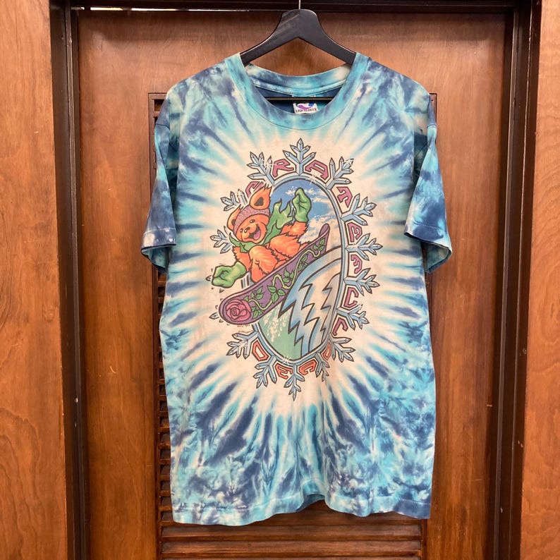 Puede incluir: Camiseta te&ntilde;ida con tinte anudado en tonos azules y blancos, con un gr&aacute;fico de un oso practicando snowboard. El oso lleva gorro y guantes, y el dise&ntilde;o est&aacute; enmarcado por un borde en forma de copo de nieve. La camiseta est&aacute; colgada en una percha negra.